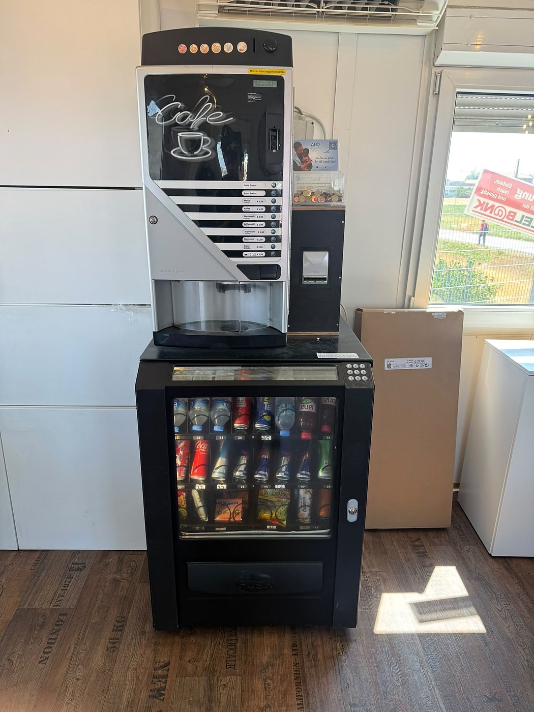 Kaffeeautomat auf einem Getränkemodul, zusammen als Kombigerät in einem hellen Raum mit Holzboden aufgestellt; im Kühlteil Wasser, Softdrinks und Riegel hinter Glas